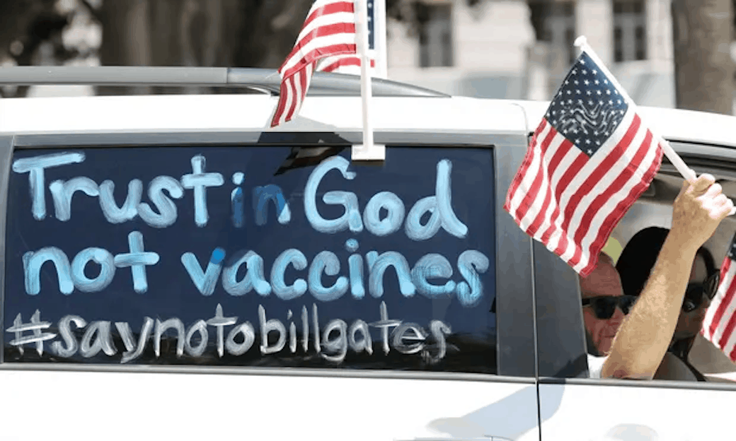 A white car draped with US flags and the words, "trust in God, not vaccines" painted on the window