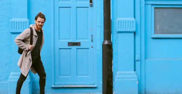 A bearded man walking towards a lampost against a blue wall in the background.