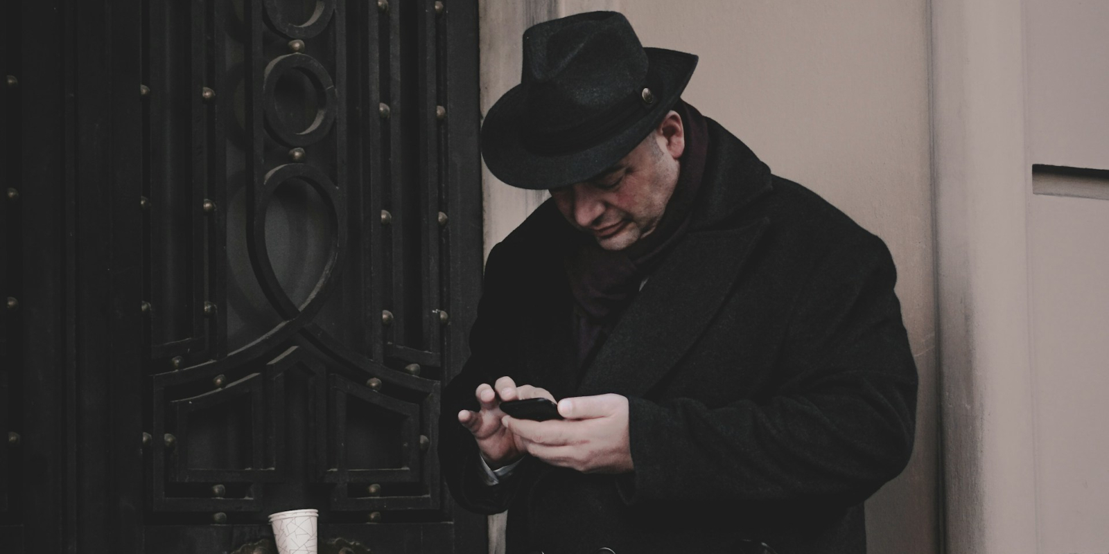 A man in a black hat and trenchcoat checking his phone in a doorway