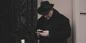 A man in a black hat and trenchcoat checking his phone in a doorway