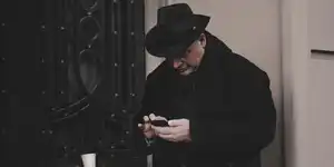 A man in a black hat and trenchcoat checking his phone in a doorway